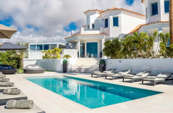 A modern villa with a pool, loungers, and landscaped surroundings under a partly cloudy sky.